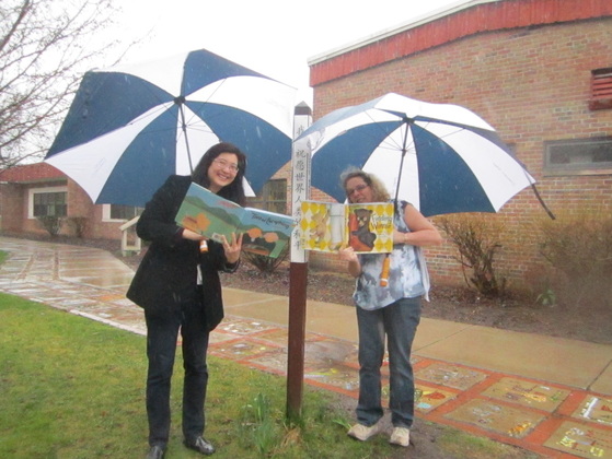 Mrs. Brown and Mrs. Bungard are outside holding an umbrella and reading a book.